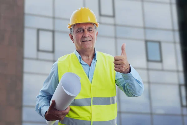 黄色のハードハットとベストを身に着けている古い男性建築家の請負業者は 仕事のコンセプトとして優しい笑顔の表現と紙の上に計画を保持ジェスチャーのように親指を作る — ストック写真