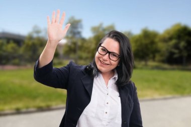 Gözlük ya da gözlük takan neşeli, yetişkin bir iş kadını ve park arka planında selamlaşmak için el sallayan şık ofis takımı.