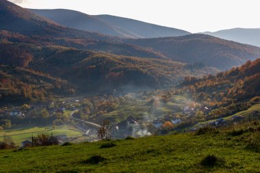 Bir dağ vadisinde küçük bir köy. Ukrayna Karpatları 'ndaki sonbahar dağ manzarası. Sarı ve kırmızı ağaçlar yeşil iğnelerle birleşmiş. Panoramik görünüm.