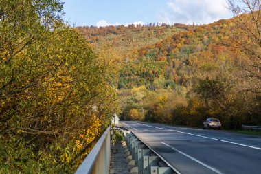 Zakarpattia Oblastı, Ukrayna - 27 Ekim 2020: Dağların arasında, ıssız bir asfalt yolun kenarında, park edilmiş bir araba var. Her yerde sonbahar ağaçları, sarı ve kırmızı yapraklar. Mavi gökyüzü.