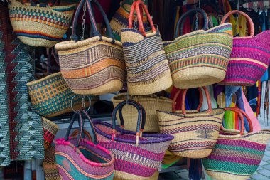 Canlı bir pazarda renkli dokuma sepetleri asılı. Çizgili desenler ve kıvrımlı saplar canlı bir zanaat gösterisi yaratıyor. Doğal iplikler ve kalın boyalar zengin zanaatkar geleneğini yansıtır.