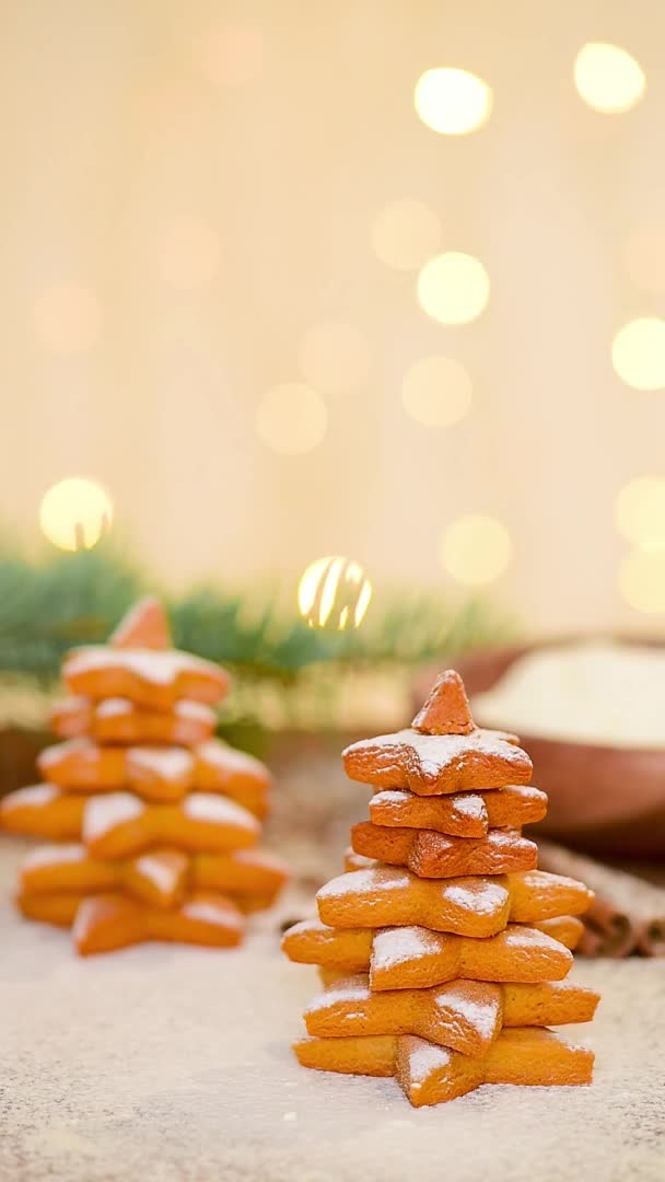 Lebkuchen in Form eines Weihnachtsbaums und eines Sterns werden mit Puderzucker gegen das Licht von Bokeh-Girlanden gestreut. Weiche Konzentration auf Lebkuchen, Zuckerschwaden. vertikales Video — Stockvideo