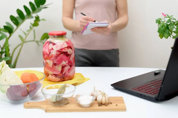 Cooking at home. a woman watches online video recipes on a laptop and cooks in the kitchen at home writes in a notebook. Pickled pink cabbage fermentative with beets and carrots in brine in a jar