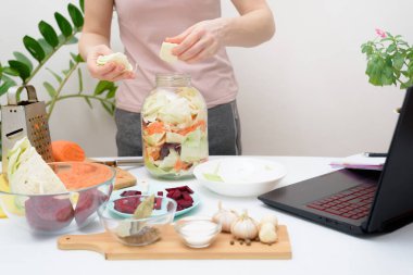 Evde yemek pişirmek. Kadın dizüstü bilgisayarda video tarifleri izliyor ve evdeki mutfakta yemek pişiriyor. Kavanozda tuzlu pancar ve havuçla birlikte salamura pembe lahana turşusu.