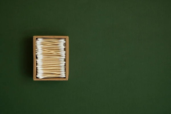 Coton-tige écologique. Une boîte en carton contenant des bourgeons de coton de bambou recyclables sur une surface verte. Concept zéro déchet. Produit écologique. Des bâtonnets de cosmétiques dans une boîte. Bâtons en bois pour nettoyer les oreilles. — Photo