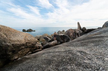 Beyaz tişörtlü, uzun saçlı genç adam bir kayanın üzerinde duruyor ve deniz manzarasının tadını çıkarıyor. Seyahat.