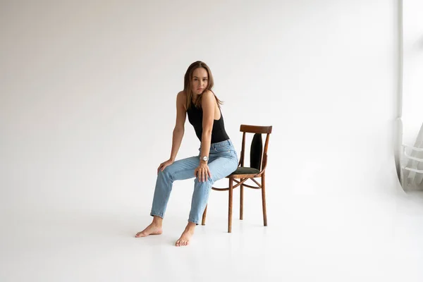 Portrait of young pensive caucasian woman posing in black underwear and blue jeans, sitting on old chair in white studio. Model tests of pretty girl in basic clothes on cyclorama. — Stock Photo, Image