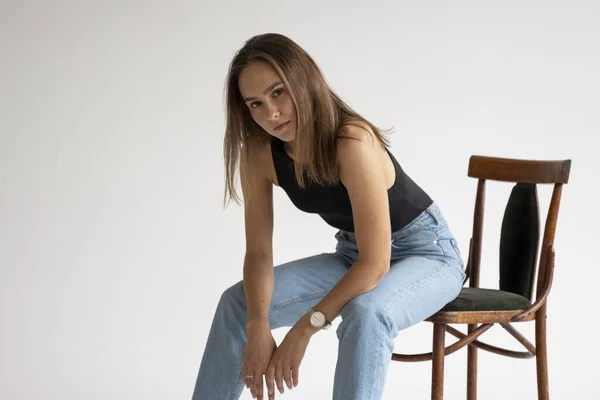Retrato de uma jovem caucasiana pensativa posando em roupa interior preta e jeans azul, sentada na velha cadeira em estúdio branco. Modelo de testes de menina bonita em roupas básicas no cyclorama. — Fotografia de Stock