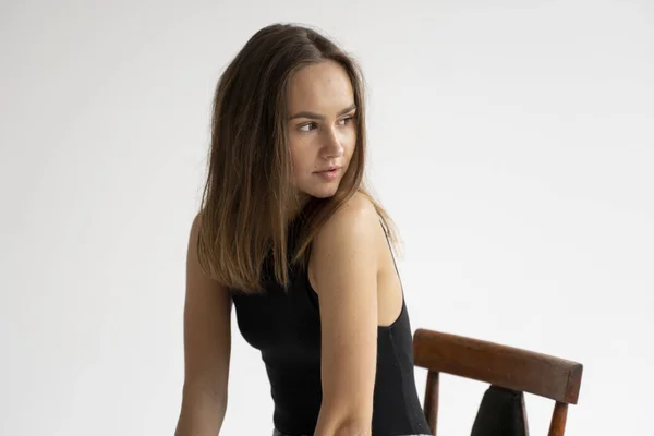 Retrato de uma jovem caucasiana pensativa posando em roupa interior preta e jeans azul, sentada na velha cadeira em estúdio branco. Modelo de testes de menina bonita em roupas básicas no cyclorama. — Fotografia de Stock