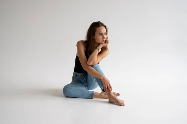 Ritratto di giovane donna felice pensierosa in posa in biancheria intima nera e jeans blu senza scarpe, seduta su un pavimento bianco in studio bianco. Modello di test di bella ragazza in abiti di base su ciclorama. — Foto Stock