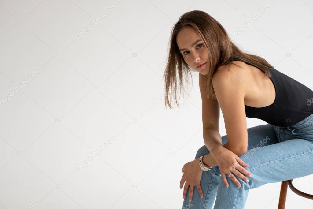 Portrait of young pensive caucasian woman posing in black underwear and blue jeans, sitting on old chair in white studio. Model tests of pretty girl in basic clothes on cyclorama.