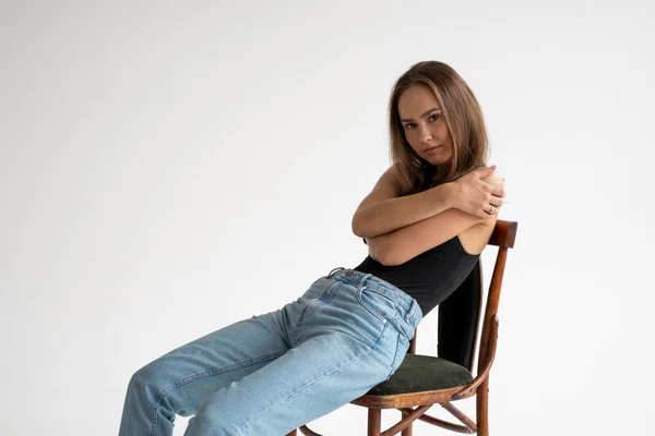 Retrato de uma jovem caucasiana pensativa posando em roupa interior preta e jeans azul, sentada na velha cadeira em estúdio branco. Modelo de testes de menina bonita em roupas básicas no cyclorama. — Fotografia de Stock