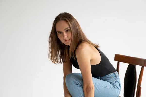 Retrato de uma jovem caucasiana pensativa posando em roupa interior preta e jeans azul, sentada na velha cadeira em estúdio branco. Modelo de testes de menina bonita em roupas básicas no cyclorama. — Fotografia de Stock