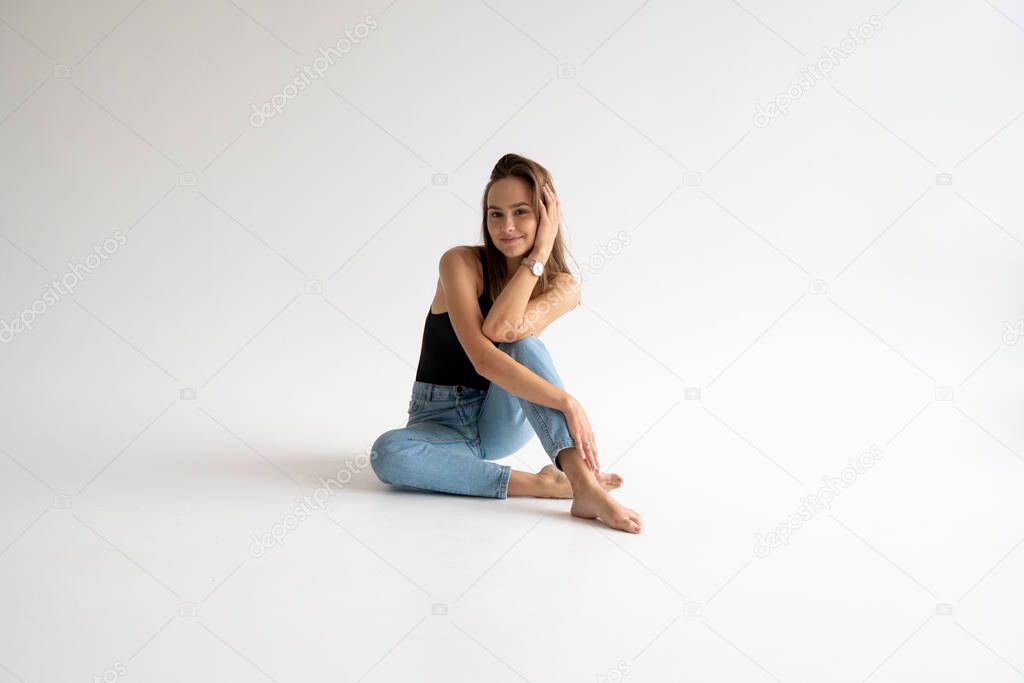 Portrait of happy young pensive woman posing in black underwear and blue jeans without a shoes, sitting on a white floor in white studio. Model tests of pretty girl in basic clothes on cyclorama.