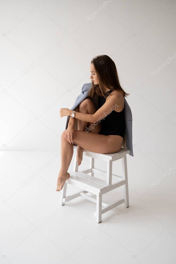 Portrait of young brunette skinny woman is posing in black underwear and blue jacket, sitting on a small white ladder in white studio. Model tests of pretty girl in basic clothes on cyclorama.