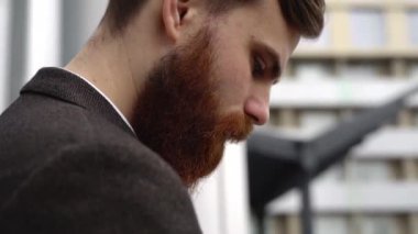 Modern genç sakallı iş adamı dijital tabletle çalışıyor. Genç hippi işadamı tableti açık havada tutuyor. Bir ofis binasının önünde dururken bir tabletle çevrimiçi çalışmak.