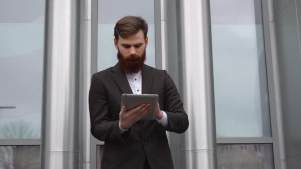Современный молодой бородатый бизнесмен работает с цифровым планшетом. Молодой бизнесмен-хипстер держит планшет в руках на открытом воздухе. Работа в Интернете с планшетом, стоя снаружи на офисном здании. — стоковое видео
