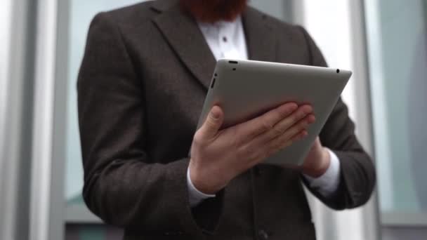 Modern Young barbudo hombre de negocios que trabaja con una tableta digital. Joven empresario hipster sosteniendo tableta en las manos al aire libre. Trabajar en línea con una tableta mientras está parado afuera en un edificio de oficinas. — Vídeos de Stock