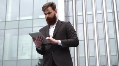 Modern genç sakallı iş adamı dijital tabletle çalışıyor. Genç hippi işadamı tableti açık havada tutuyor. Bir ofis binasının önünde dururken bir tabletle çevrimiçi çalışmak.