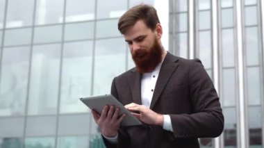 Modern genç sakallı iş adamı dijital tabletle çalışıyor. Genç hippi işadamı tableti açık havada tutuyor. Bir ofis binasının önünde dururken bir tabletle çevrimiçi çalışmak.