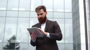 Modern genç sakallı iş adamı dijital tabletle çalışıyor. Genç hippi işadamı tableti açık havada tutuyor. Bir ofis binasının önünde dururken bir tabletle çevrimiçi çalışmak.
