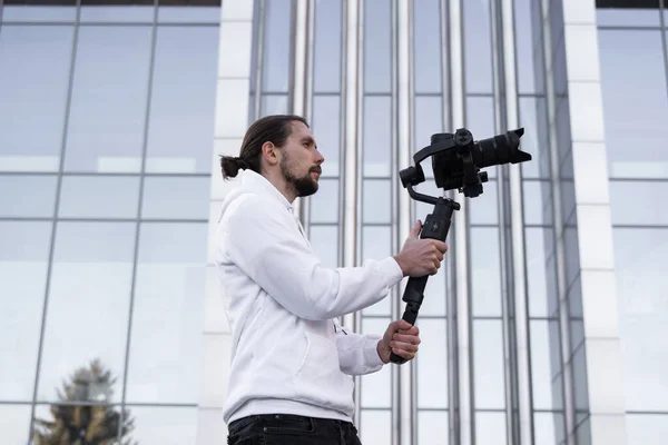 Giovane videografo professionale con fotocamera professionale su stabilizzatore gimbale a 3 assi. L'attrezzatura Pro aiuta a fare video di alta qualità senza scuotere. Cameraman indossa felpa bianca facendo un video . — Foto Stock