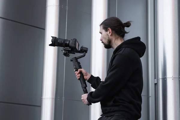 Ung professionell videograf som håller professionell kamera på 3-axlig gimbal stabilisator. Pro-utrustning hjälper till att göra högkvalitativ video utan att skaka. Kameraman bär vit luvtröja gör en video. — Stockfoto