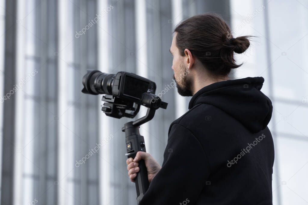 Young Professional videographer holding professional camera on 3-axis gimbal stabilizer. Pro equipment helps to make high quality video without shaking. Cameraman wearing white hoodie making a videos.