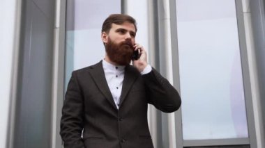 Finans bölgesinde iş adamı telefonda konuşuyor. Ofis binasının üzerinde akıllı telefon kullanan genç bir şehirli adamın portresi. İş adamı dışarıda akıllı telefonuyla konuşuyor. Teknoloji.