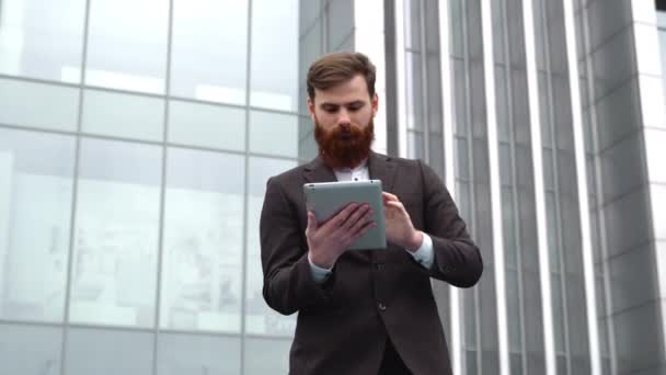 Modern genç sakallı iş adamı dijital tabletle çalışıyor. Genç hippi işadamı tableti açık havada tutuyor. Bir ofis binasının önünde dururken bir tabletle çevrimiçi çalışmak. — Stok video
