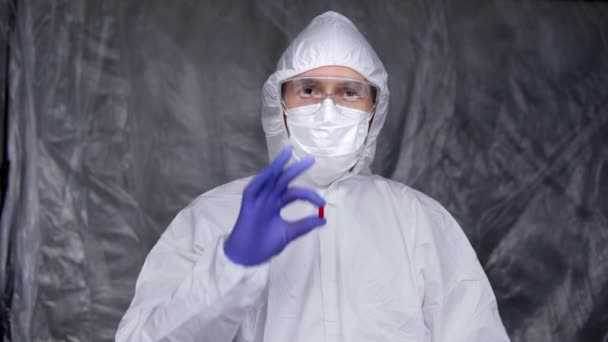 El médico con traje protector blanco, máscara médica y guantes de goma sostiene una píldora roja como un medicamento contra la amenaza pandémica del coronavirus. Epidemia, pandemia de coronavirus covid 19. — Vídeos de Stock