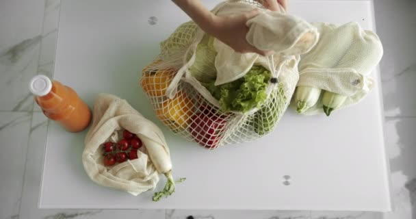 Vrouwen met een herbruikbare boodschappentas met groenten op een witte schone keuken thuis na het boodschappen doen. Geen afval en plastic vrij concept. Mesh katoenen shopper met groenten. Ecologie. — Stockvideo