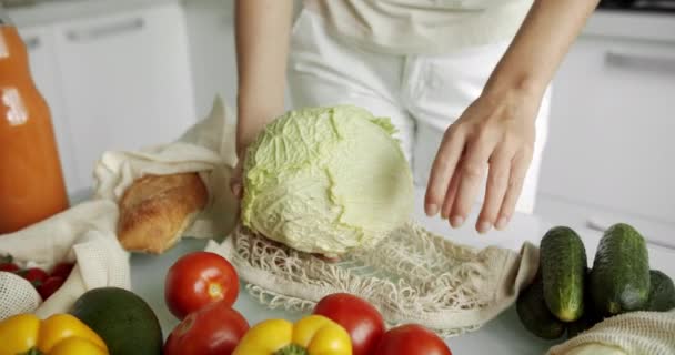 Mujer poner una col dentro de la bolsa de comestibles reutilizables con verduras en una mesa con pimientos, tomates y pepinos en la cocina. Residuos cero, plástico gratis, compras de comestibles. Comprador de algodón de malla. — Vídeos de Stock