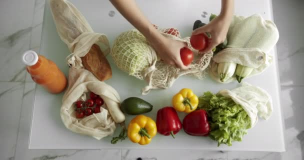 La mujer saca los tomates de la bolsa de la compra reutilizable con hortalizas en la mesa en la cocina en casa después de la compra. Cero residuos y concepto libre de plástico. Comprador de algodón de malla. — Vídeos de Stock