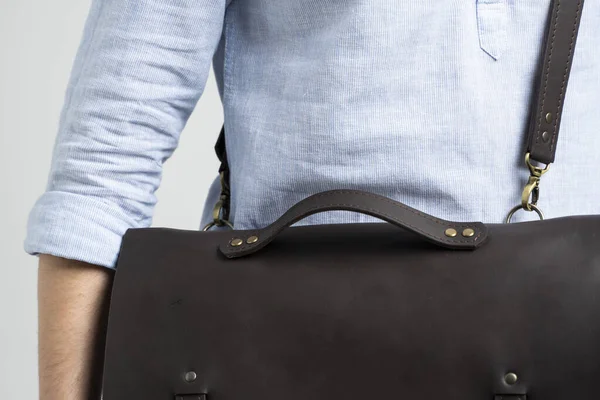 Детали коричневой мужской плечевой кожаной сумки для документов и ноутбука на плечах мужчины в синей рубашке и джинсах с белым фоном. Сумка, мужской кожаный портфель ручной работы. — стоковое фото