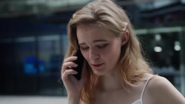 Young business woman in a white t-shirt is laughs and talking on the phone with business partners or friends with a business center or shopping mall behind. Happy girl tells or hears something funny. — Stock Video