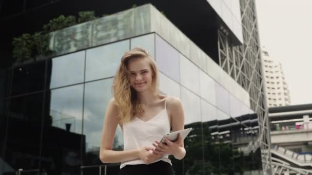 Портрет бізнес-леді в білій футболці використовує планшетний комп'ютер з бізнес-центром позаду, на відкритому повітрі. Ділова жінка на зустрічі і чекає ділового партнера і тримає планшет в руках . — стокове відео