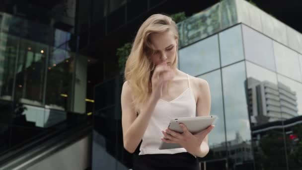 Porträtt av affärskvinna i vit t-shirt använder en surfplatta med ett businesscenter bakom, utomhus. Affärskvinna på möte och väntande affärspartner och hålla en surfplatta i händerna. — Stockvideo