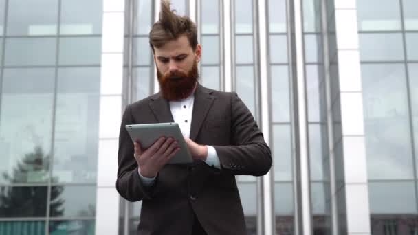 Modern Young barbudo hombre de negocios que trabaja con una tableta digital. Joven empresario hipster sosteniendo tableta en las manos al aire libre. Trabajar en línea con una tableta mientras está parado afuera en un edificio de oficinas. — Vídeos de Stock