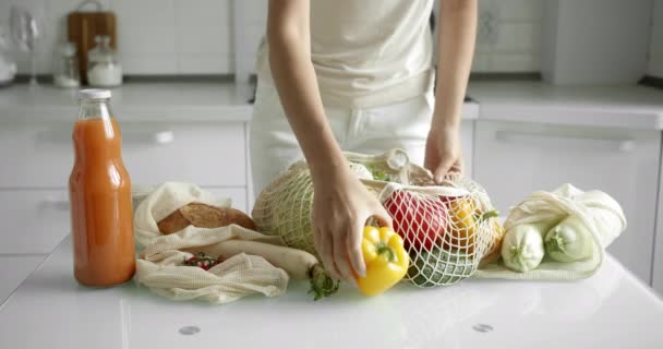 Vrouw neemt gele en rode peper uit herbruikbare boodschappentas met groenten op een tafel in de keuken thuis na boodschappen doen. Geen afval en plastic vrij concept. Mesh katoenshopper. — Stockvideo