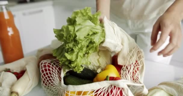 Vrouw haalt salade uit herbruikbare kruidenierszakje met groenten op een tafel met paprika 's, tomaten en komkommers in de keuken. Geen afval, plastic vrij concept, boodschappen doen. Mesh katoenshopper. — Stockvideo