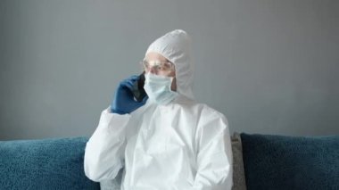Koruyucu beyaz takım elbiseli ve tıbbi maskeli bir adam Coronavirüs salgını yüzünden evinde yatağında oturan bir telefonu kullanıyor. Salgın sırasında uzaktan çalışma. COVID-19 karantina sırasında evde kal.