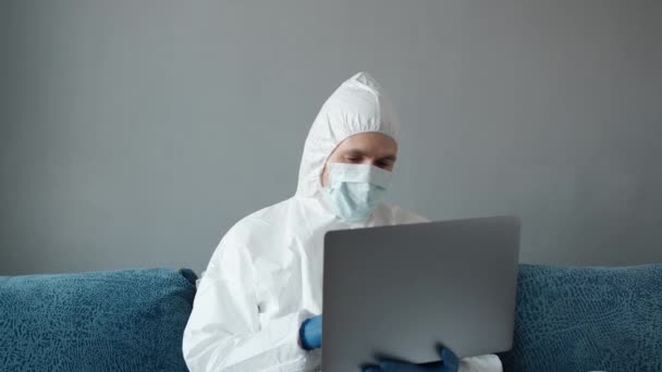 Mannen i skyddande vit kostym och medicinsk mask arbetar hemifrån på en soffa med laptop på grund av coronavirus pandemi. Fjärrarbete under en pandemi. Stanna hemma under COVID-19 karantänkoncept. — Stockvideo
