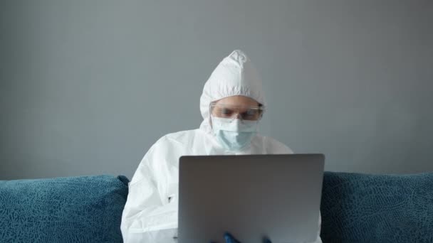 Άνδρας με προστατευτική λευκή στολή και ιατρική μάσκα εργάζεται από το σπίτι σε έναν καναπέ με φορητό υπολογιστή λόγω της πανδημίας του κορωναϊού. Απομακρυσμένη εργασία κατά την πανδημία. Μείνετε στο σπίτι κατά τη διάρκεια COVID-19 καραντίνα έννοια. — Αρχείο Βίντεο