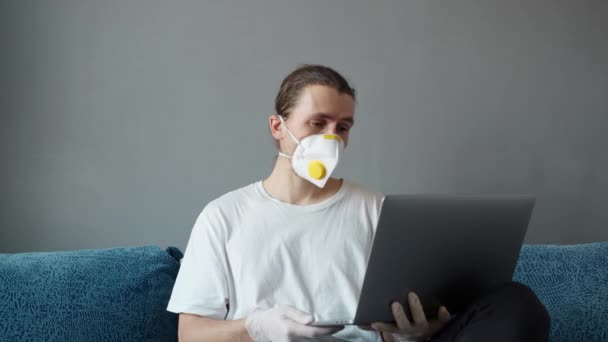 Man i t-shirt och medicinsk mask, respirator arbetar på distans hemma på en soffa med bärbar dator talar med vänner eller partners via webbkamera. Coronavirus pandemi. Stanna hemma, COVID-19, karantän. — Stockvideo