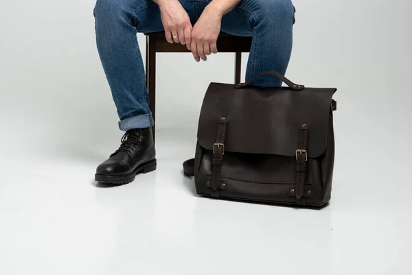 Mężczyzna w niebieskich dżinsach i czarnych butach siedzi na krześle z brązową męską torbą na ramię na dokumenty i laptopem na białej podłodze. Torba męska skórzana, torby kurierskie, ręcznie robiona teczka. — Zdjęcie stockowe