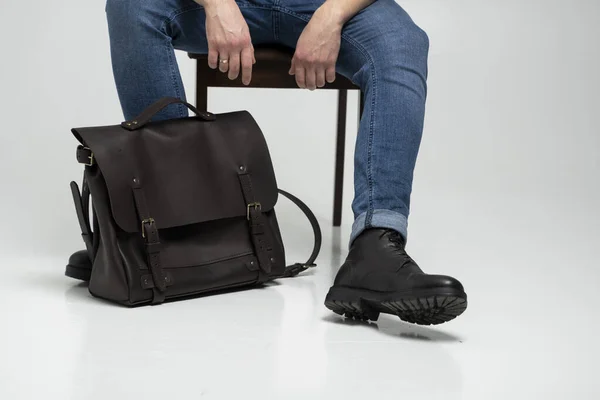 El hombre en pantalones vaqueros azules y botas negras se sienta en una silla con un bolso de cuero marrón para hombre para documentos y un portátil en un piso blanco. Bolso de cuero para hombre, bolsos de mensajero, maletín hecho a mano. — Foto de Stock