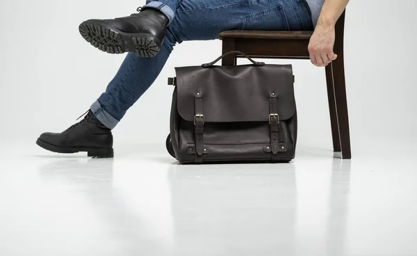 Mężczyzna w niebieskich dżinsach i czarnych butach siedzi na krześle z brązową męską torbą na ramię na dokumenty i laptopem na białej podłodze. Torba męska skórzana, torby kurierskie, ręcznie robiona teczka. — Zdjęcie stockowe