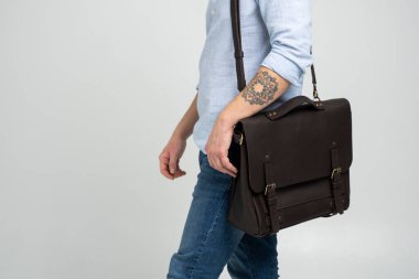 Kahverengi erkek omuz çantası bir belge ve dizüstü bilgisayar için mavi gömlekli ve beyaz arka planlı kot pantolonlu bir adamın omuzlarında. Çanta, erkek derisi el yapımı çanta..