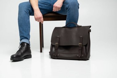 Mavi kotlu ve siyah çizmeli bir adam, kahverengi erkek omuz çantalı bir sandalyede oturuyor. Belgeler için ve dizüstü bilgisayar için beyaz bir katta. Erkek derisi çanta, kurye çantası, el yapımı çanta..